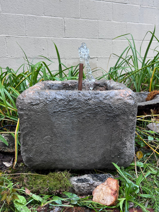 Decorative stone with water feature surrounded by grass and plants on a concrete surface