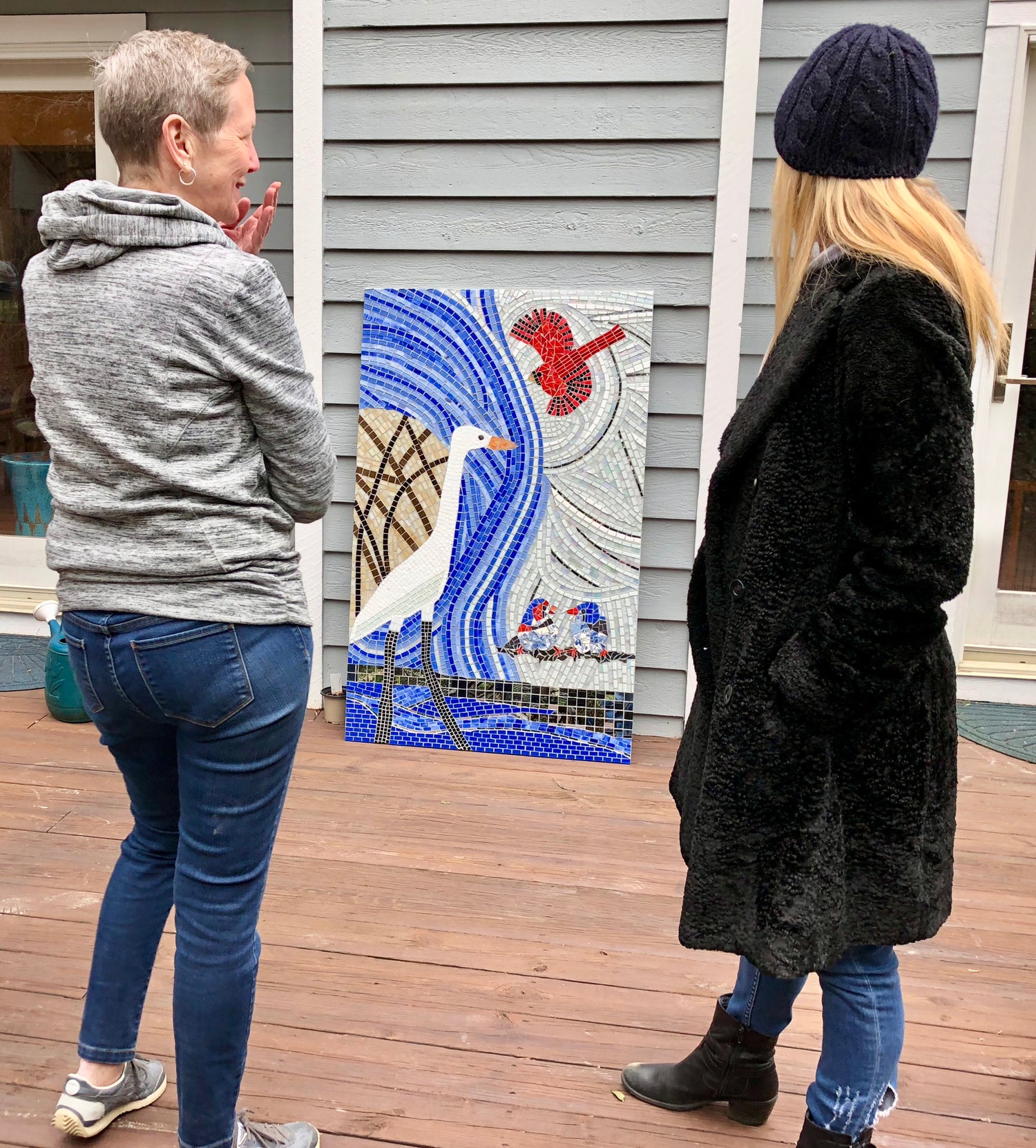Two people standing on a wooden deck looking at a mosaic artwork featuring birds.